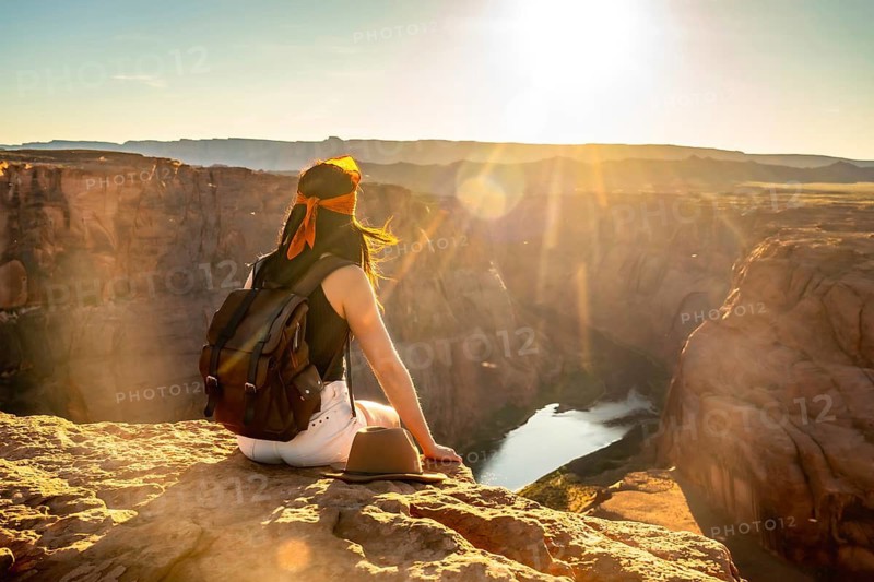 Woman backpacker sitting on cliff edge admiring the vast canyon landscape and colorado river during a golden hour sunset, representing adventure, travel, and freedom