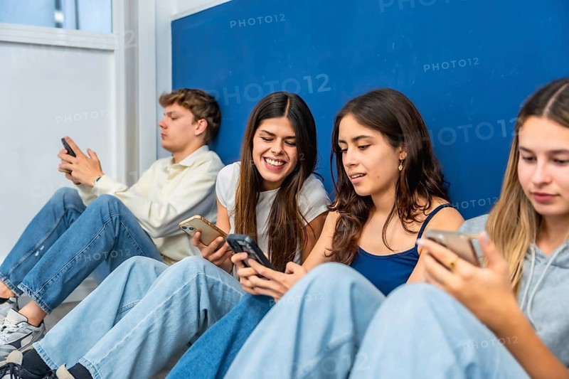 Young diverse students sitting on the floor, some laughing, while interacting and absorbed by their mobile phones against a blue and white wall, representing technology use in youth