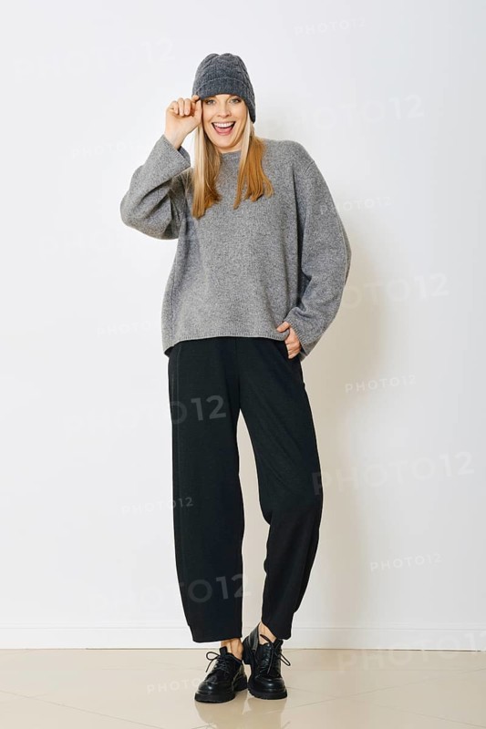 Laughing woman in gray sweater and black pants touching knitted hat. She smiles and lifts her hand playfully