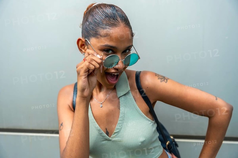 Young woman confidently pulling down round sunglasses looking at the camera with an open mouth and joyful expression against a minimalist urban background