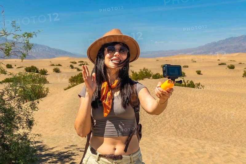 Woman wearing a hat and sunglasses, vlogging her desert adventure for social media, holding an action camera on a selfie stick against the vast death valley mesquite flat sand dunes landscape