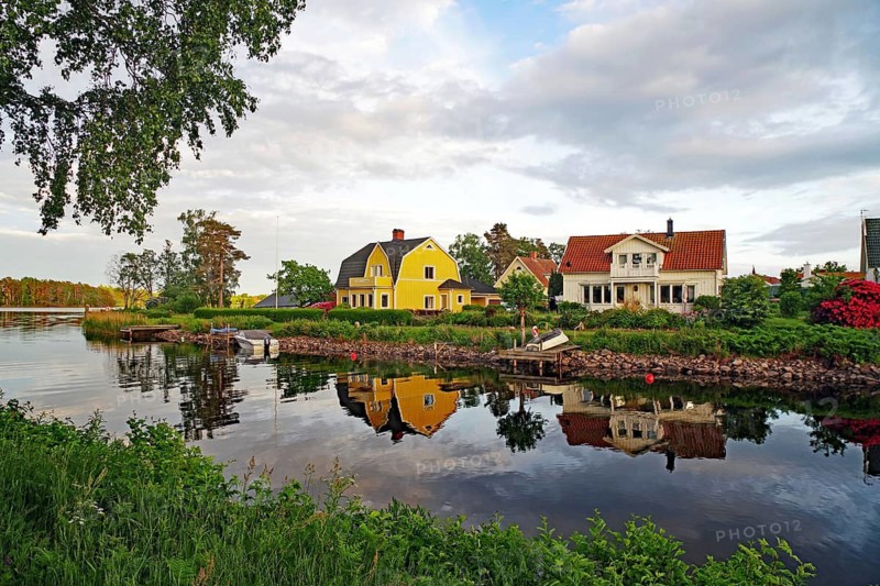 Two charming houses with clear reflections in the river, surrounded by lush nature and cloudy sky, Karlsborg, Västra Götalands län, Sweden