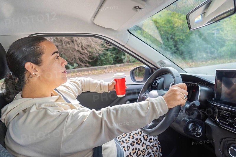 Woman driving with one hand on steering wheel while holding red coffee cup, profile view, car interior, road trip concept