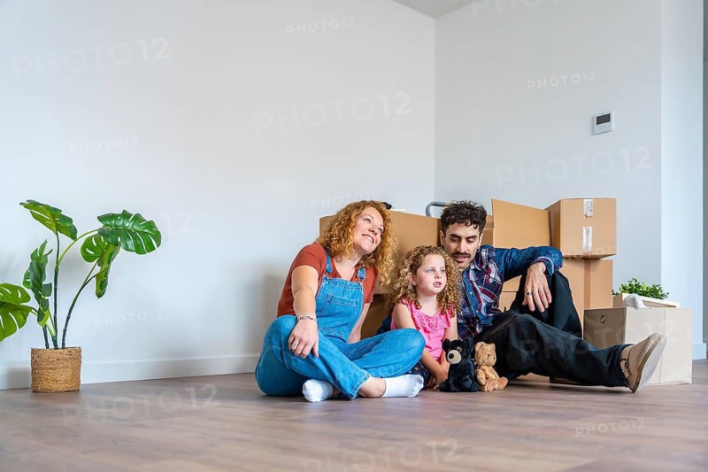 Happy family with parents and daughter sitting on floor in empty living room, surrounded by cardboard boxes, moving into new house and planning their future