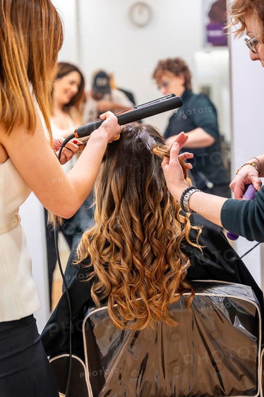 Professional hairdressers styling a client's long brown and blonde ombre hair with a flat iron, creating beautiful curls for a fresh look in a modern beauty salon
