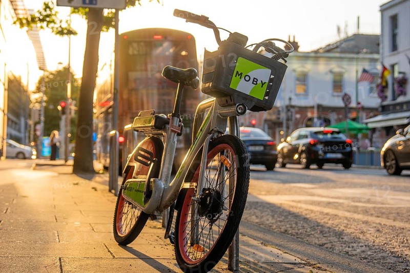 A Moby rental bike is parked on the side of the road at sunset, traffic moving in the background, Dublin, Ireland