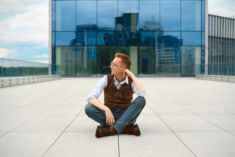 Middle-aged man sits cross-legged on rooftop in urban area, looking thoughtfully to side. Tall buildings surround him. Bright daylight shines down, revealing clear sky