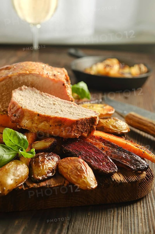 Slice of roasted pork on wooden cutting board beside roasted vegetables and green herbs. A glass of white wine is in background. Candlelight adds warmth to dinner table setting at evening