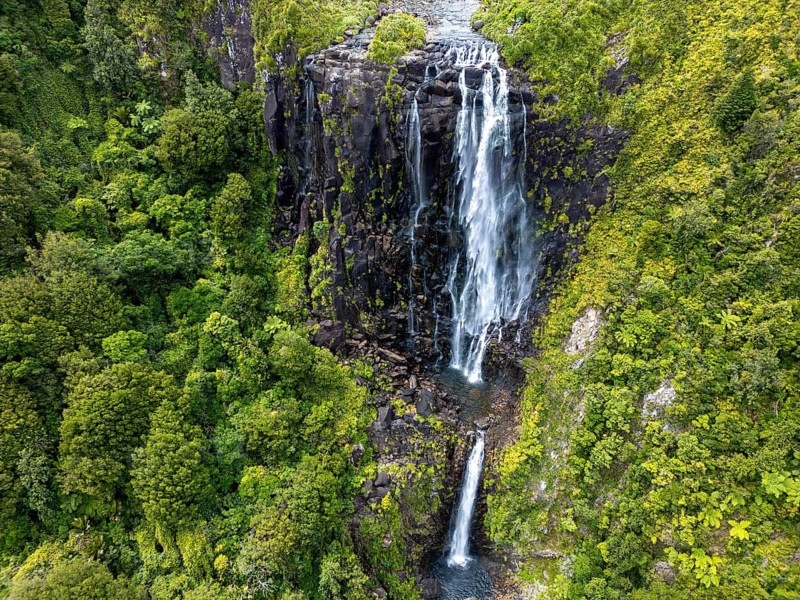 Stunning aerial photo of a tall waterfall flowing down dark volcanic rock surrounded by lush green forest. Captured near Rotorua, New Zealand, in a geothermally active region