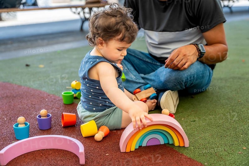Young child creating a rainbow arch with educational wooden blocks while father watches, enjoying a joyful moment of learning and playtime outdoors on a soft playground surface