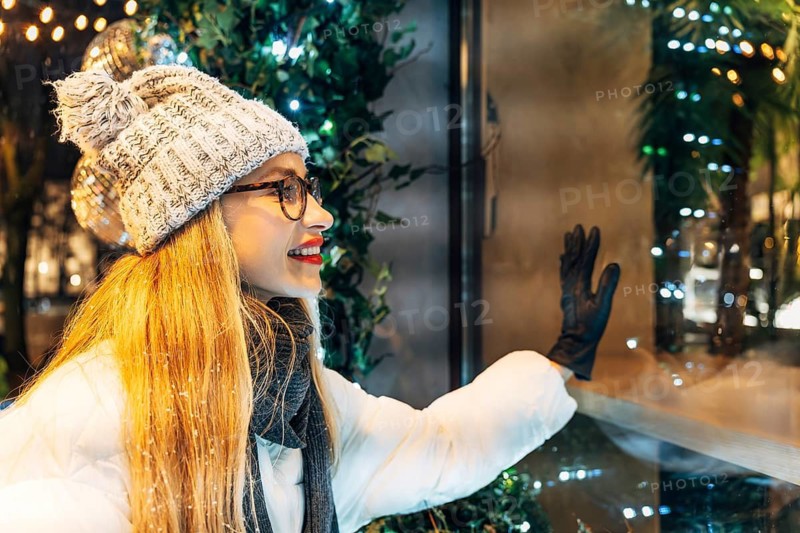 Happy young woman in winter clothing looking at store window display. A woman wearing a white winter coat, gray knit hat with a pom-pom, black gloves and glasses stands beside a glass window decorated with warm holiday lights and greenery. Her gloved hand touches the reflective surface, capturing soft highlights from the surrounding illumination. The scene combines winter attire, glowing seasonal decorations and night reflections, creating a cozy festive atmosphere
