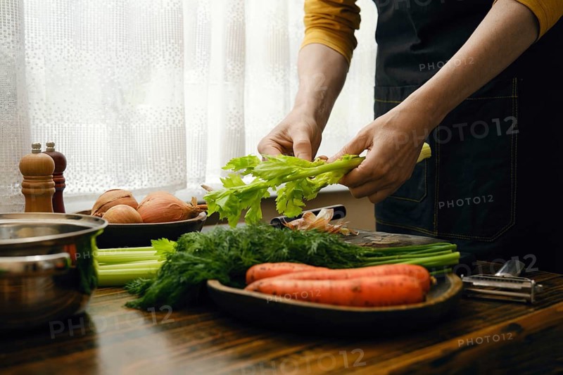 Person tears off a celery stalk. Other vegetables like carrots and onions are nearby. Light comes through the window