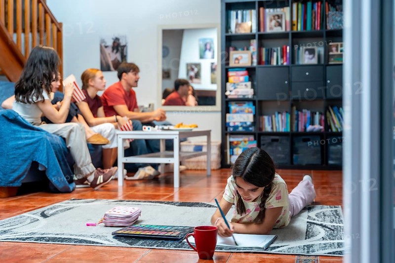 Family members relaxing in their living room, an adult couple and two teenagers sitting on a sofa while a young girl lies on the floor drawing and coloring, enjoying leisurely time