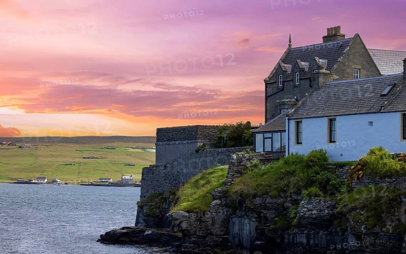 Historic stone buildings perched on dramatic rocky cliffs overlooking the sea at vivid pink and orange sunset in Lerwick, Shetland Islands, Scotland, United Kingdom
