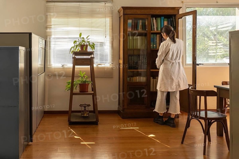 Rear view of a young female scientist in white lab coat standing in front of an open antique wooden bookcase in a bright sunlit office with indoor plants and wooden floor