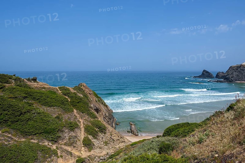 Rocky cliffs with green vegetation and wavy sea under a clear blue sky, Odeceixe, Faro, Portugal