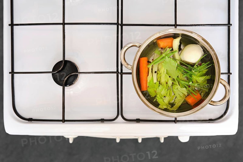 The process of cooking vegetable broth. A pot on a stove with celery, carrot, onion, and other vegetables inside it. The pot is full of water and is heating up