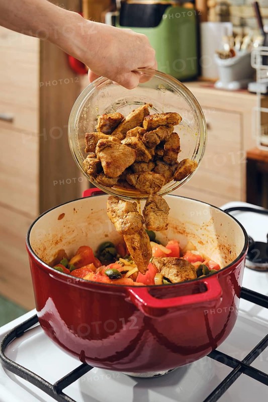 Person is pouring fried pork pieces into pot filled with fresh vegetables on stove. Process of making meat and vegetable stew
