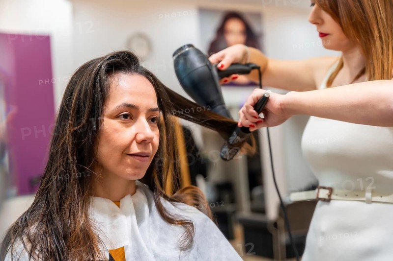 Female hairdresser drying and styling a customer's long hair with a hairdryer and brush, providing professional beauty service in a modern hair salon environment