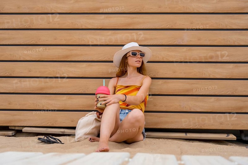 Woman wearing a straw hat and sunglasses, sitting barefoot on a sandy beach next to a wooden wall, holding a refreshing smoothie, enjoying summer vacation and leisure