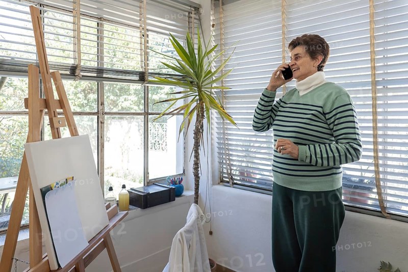 Wide shot of an older woman in striped sweater smiling during a phone call in her home studio, with easel, yucca plant, large windows and art supplies all visible around her