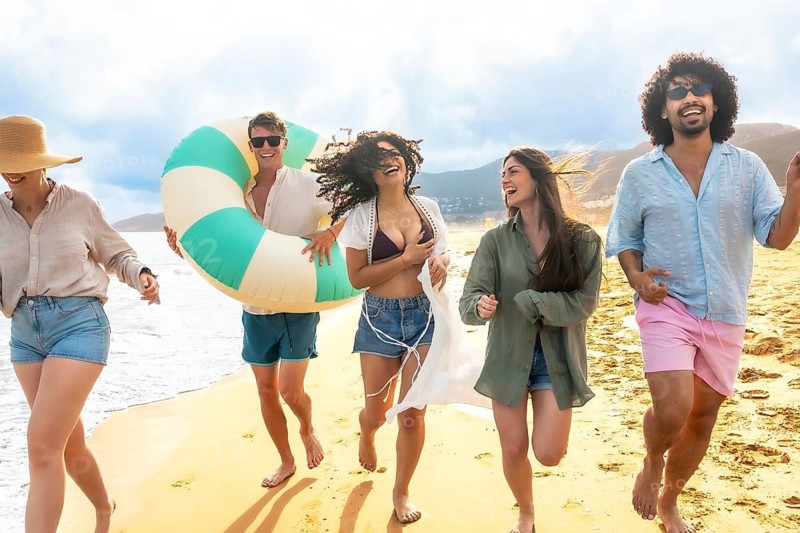 Group of diverse friends running barefoot along the sandy beach, carrying an inflatable ring and laughing together during a joyous summer vacation by the sea