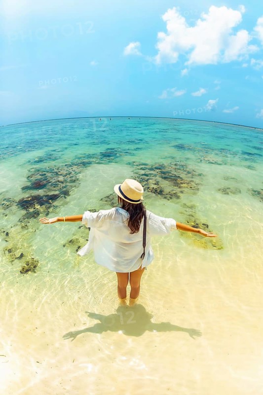 Young carefree woman wear straw hat stands arms wide open hug ocean in shallow, crystal-clear turquoise water on a vast tropical beach. Fish eye lens captures a panoramic view of coral reefs white sand