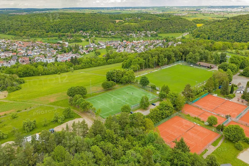 Aerial view of a sports complex with a green soccer field and several red tennis courts, surrounded by lush countryside and a village, Aidlingen sports field, Böblingen district, Germany