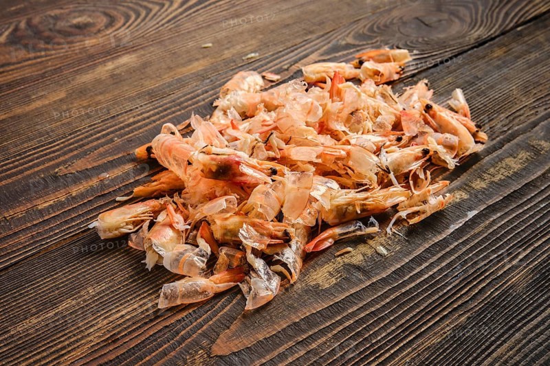 Shrimp shells are piled on a clean wooden table. They come from a recent seafood meal preparation. The setting shows the aftermath of peeling shrimp for cooking