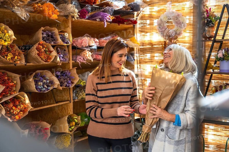 Florist smiling while assisting an older female customer picking out a wrapped dried flower bouquet, reflecting successful small business retail with happy client interaction