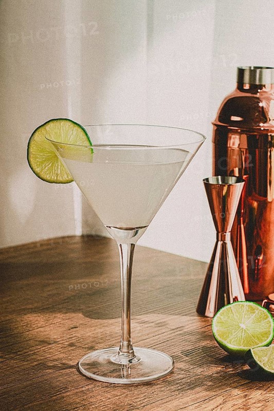 A clear cocktail sits in a glass with a lime slice on the rim. A copper shaker and halved lime are also on the wooden table. Bright light comes in from the window