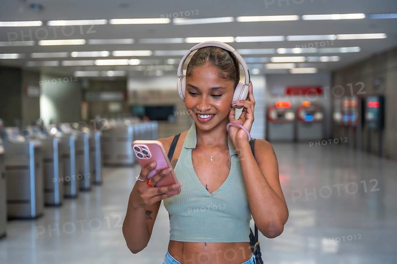 Young woman smiling while listening to music with headphones and using a smartphone, navigating modern technology and entertainment in a bustling public transit environment