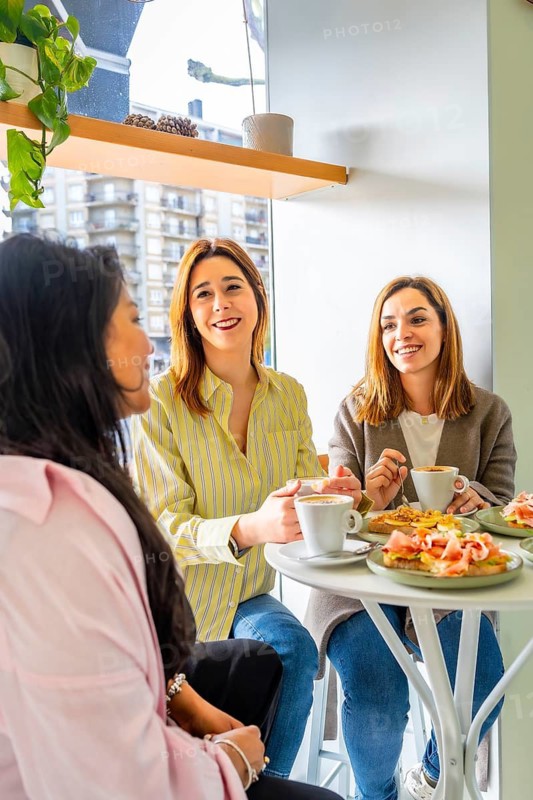 Three young women smiling and chatting over coffee and brunch at a sunny cafe window table, enjoying food, conversation and relaxed, multicultural weekend togetherness