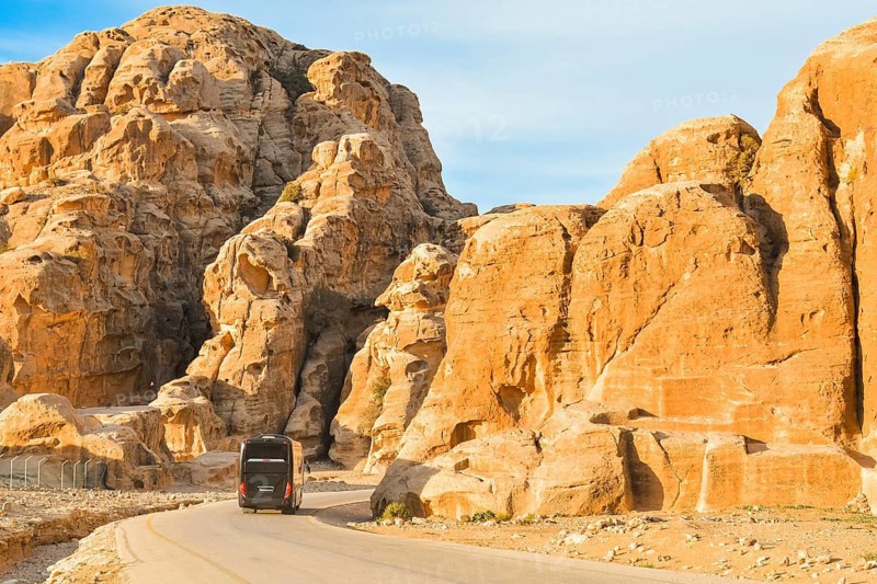 Black passenger coach bus driving asphalt road winding through sandstone rock mountains canyon desert landscape leading ancient archaeological tourist attraction Little Petra Jordan