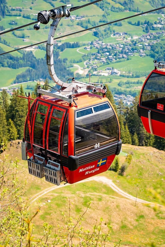 Red cable car cabin floating over green landscape with a view of a village in the distance, Hahnenkamm, Kitzbühel, Austria