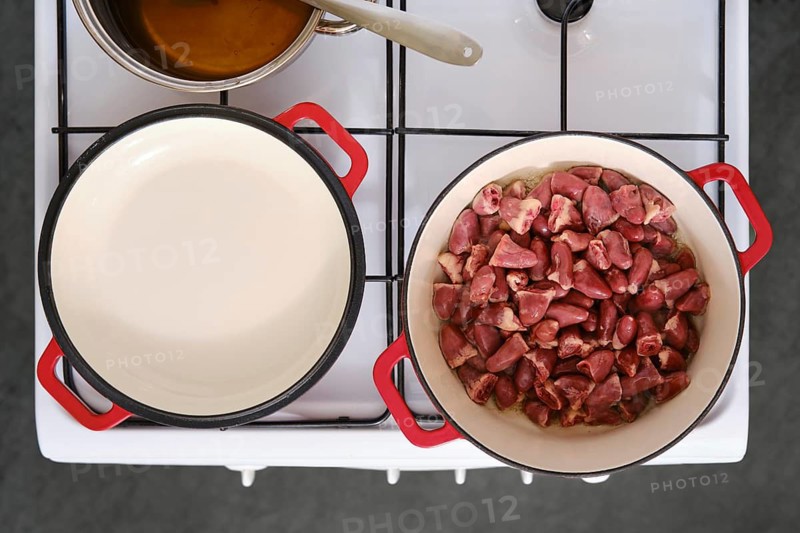 A stove is set up in a kitchen with two pots. One pot has raw chicken hearts, while the other pot contains a sauce. Cooking tools are placed nearby, ready for use