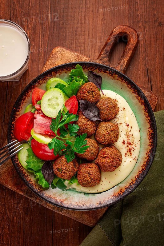 A plate holds falafel balls, tahini, salad with tomatoes and cucumbers, and a drink. The setting is warm and inviting, perfect for a midday meal with fresh ingredients