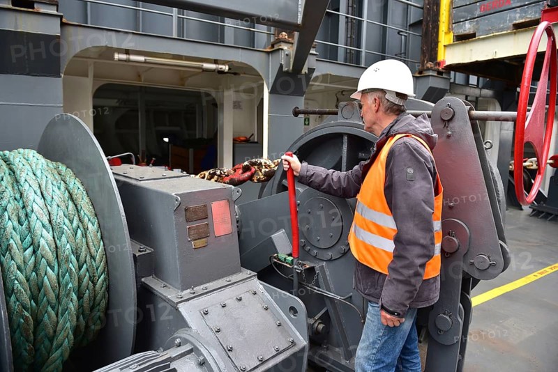 A worker with helmet and vest handles a rope on a large machine on a ship, worker with the anchor chain on a container ship, Schleswig-Holstein, Germany