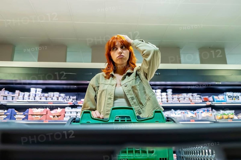 Young woman standing in a supermarket aisle, looking thoughtful and concerned about inflation and increasing grocery costs while hands resting on a green shopping cart