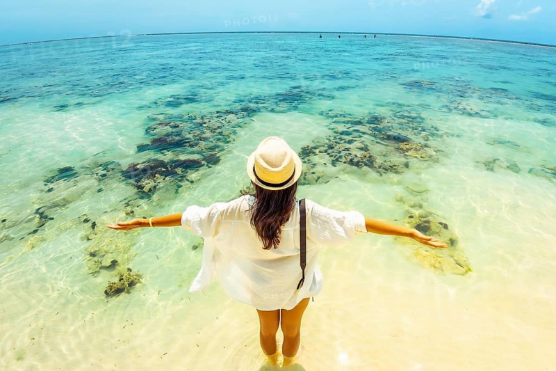 Young carefree woman wear straw hat stands arms wide open hug ocean in shallow, crystal-clear turquoise water on a vast tropical beach. Fish eye lens captures a panoramic view of coral reefs white sand