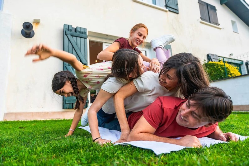 Multi generational family members bonding while creating a playful pile up on the green grass in a home garden, sharing laughter and enjoying togetherness outdoors