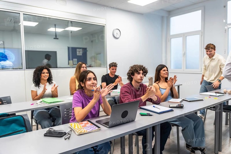 Happy young university students clapping and smiling during a lecture or seminar, engaged and celebratory in a modern classroom with laptops, notebooks, and bright interior