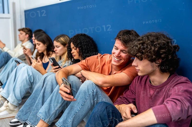 Diverse group of young students sitting in a row, individually focused on their mobile phones while one smiling man engages with another, highlighting digital communication and social connection