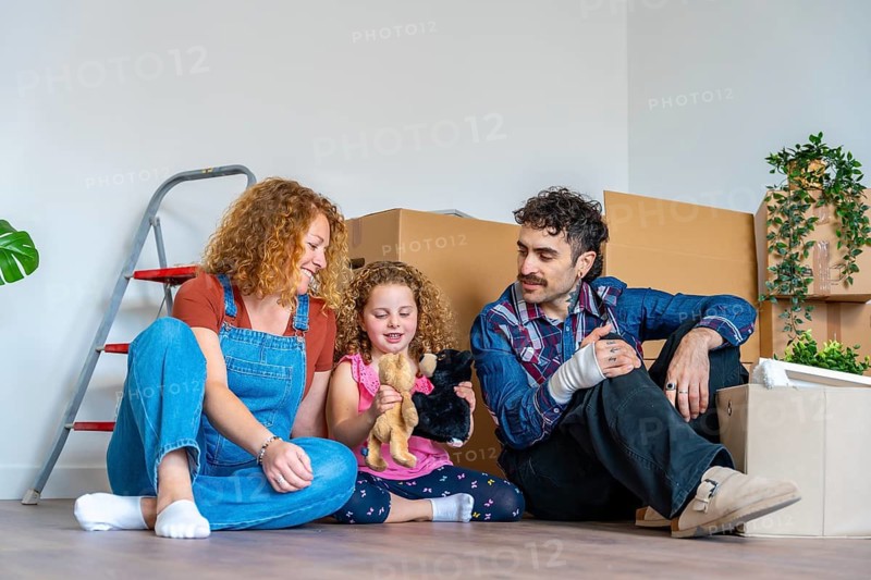 Happy family parents and daughter sitting on the floor amid moving boxes, smiling and celebrating their fresh start in a new home, unpacking, relaxed and joyful together