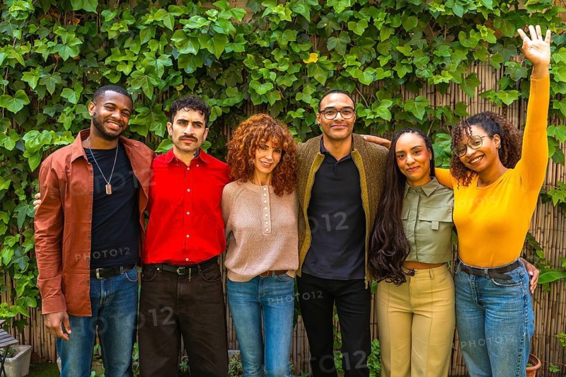 Diverse group of smiling adults supporting each other, standing together outside against a green leaf wall, representing concepts of community, wellbeing, and mental health