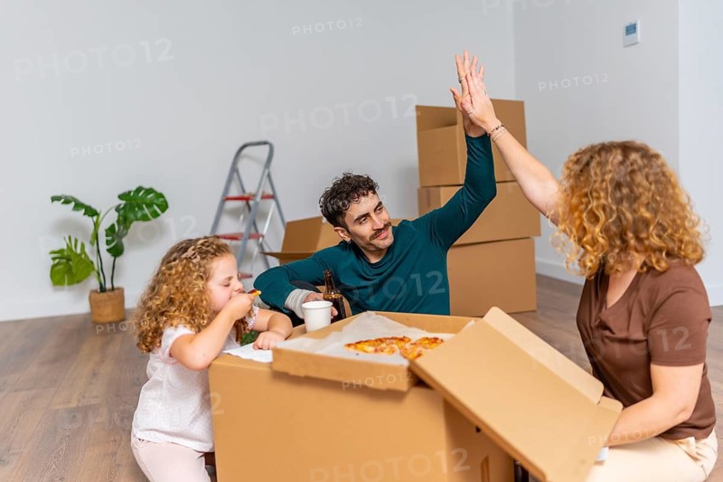 Family celebrating moving day in their new house, giving a high five while eating pizza on cardboard boxes, symbolizing relocation and a happy fresh start