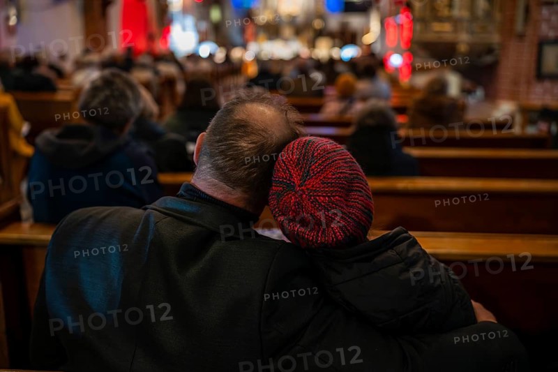 View from behind of a couple sitting closely in wooden church pews during a service, with the woman wearing a red knitted hat and leaning on his shoulder in Lubin, Poland