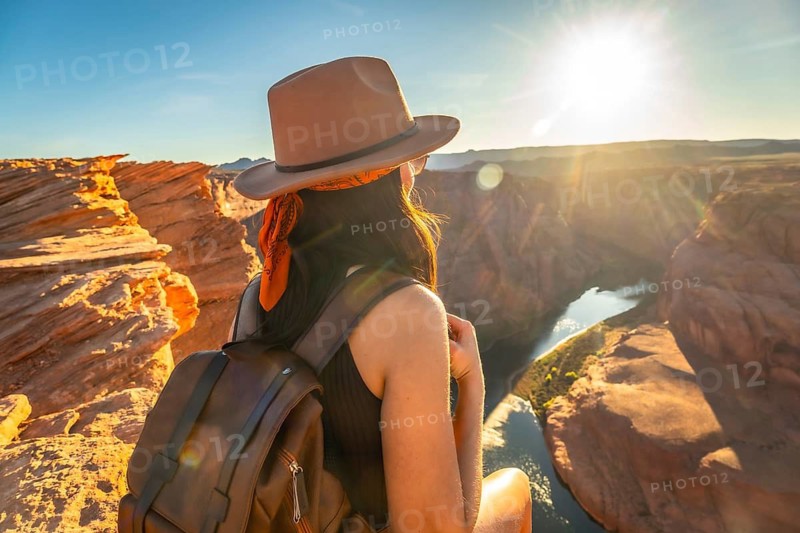 Woman wearing a hat and backpack sitting on a cliff edge, looking at the sunrise over the colorado river making a famous u turn creating a stunning desert landscape