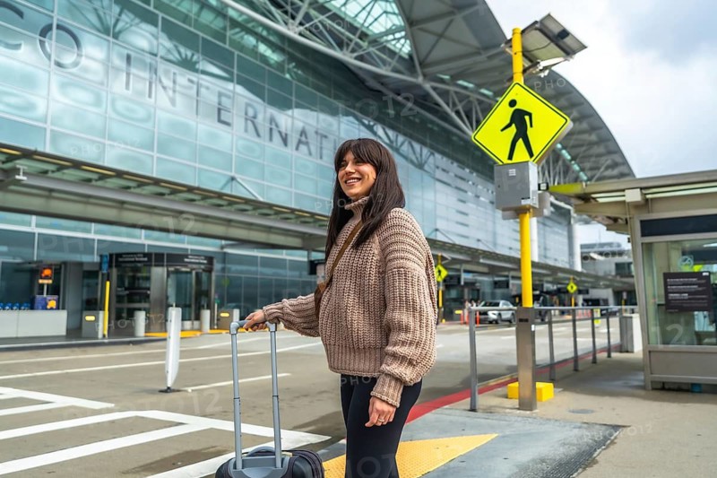 Young woman smiling while pulling her hardshell carry on luggage across an airport crosswalk, arriving for her flight or departing after a journey, focusing on travel and transportation