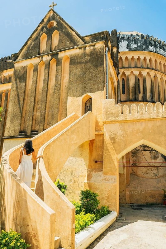 Caucasian Woman tourist wear white dress explores visit historic Christ Church Anglican Cathedral in Stone Town, Zanzibar. Features unique Gothic and Arabic coral stone in sunny day architecture